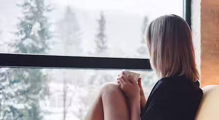 Photo of girl sitting inside home looking out window at snow outside