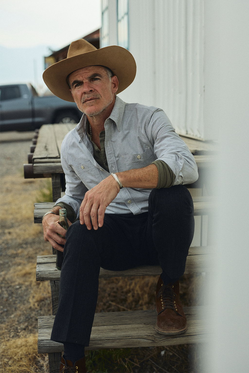 Micahel Kelly at an abandoned service station in Livingston, Montana drinking a Coke wearing the Bridges Oxford Workshirt in Olive and the Rivay Italian Wool Suit in Charcoal Herringbone Trousers