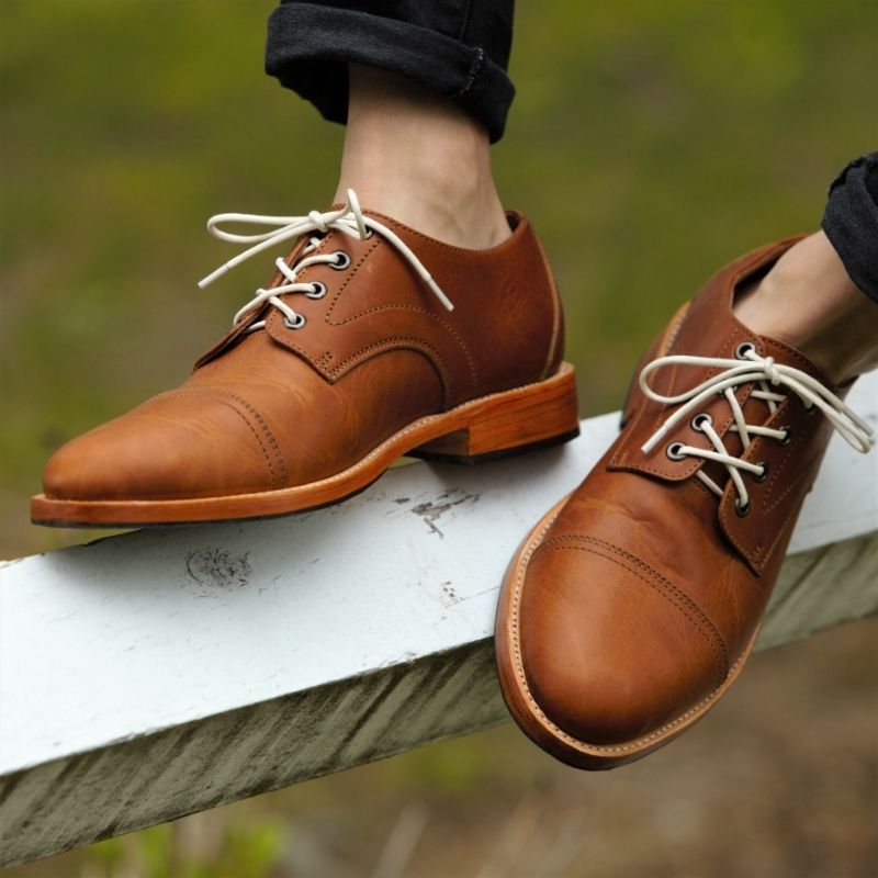 An outdoor lifestyle shot from the ankles down of a person resting their feet on a weathered white wooden fence rail. They are wearing light brown or caramel leather Adelante Marco cap-toe derby shoes with contrasting cream-colored laces and a stacked leather sole. The look is styled with rolled-up black pants against a softly blurred green grass background.
