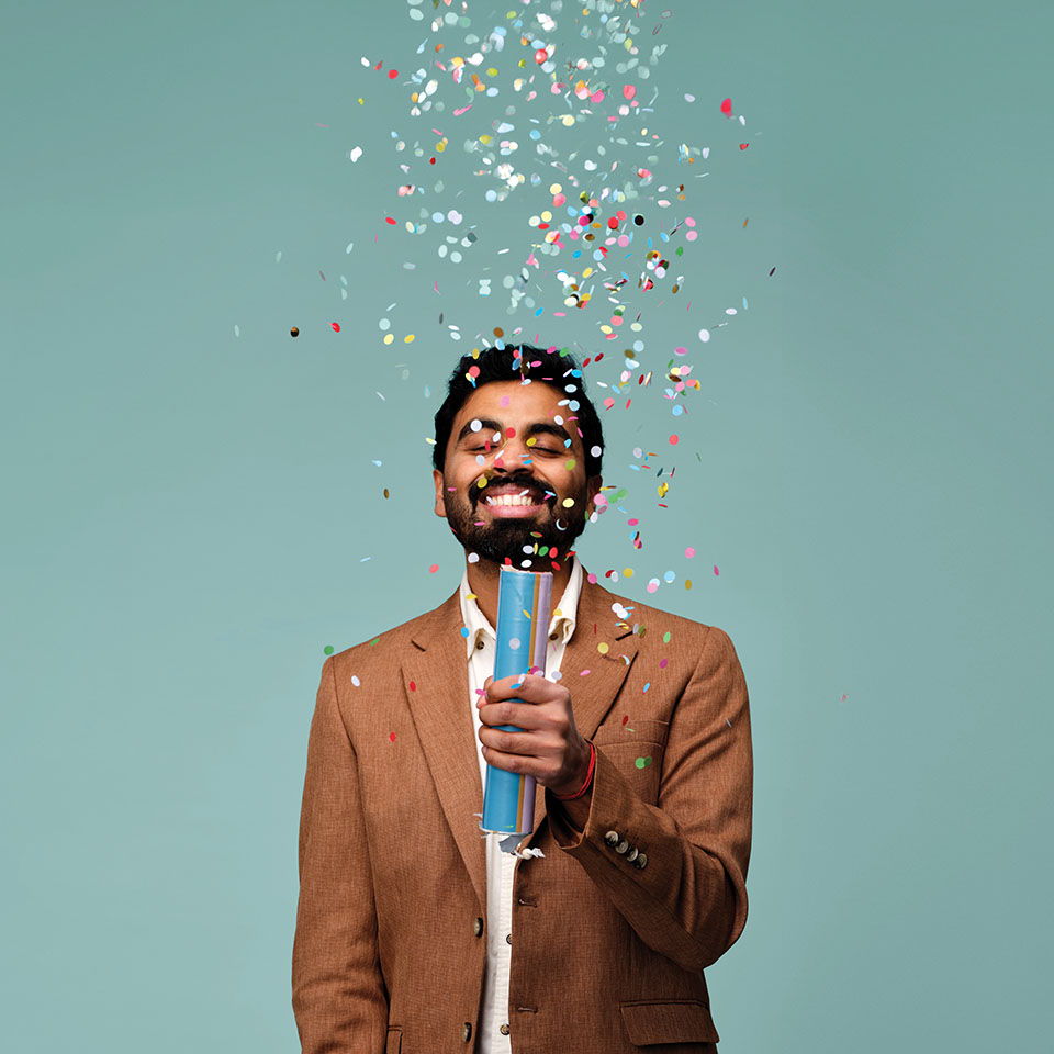 Man in a brown jacket holding a confetti popper, confetti falling around him.