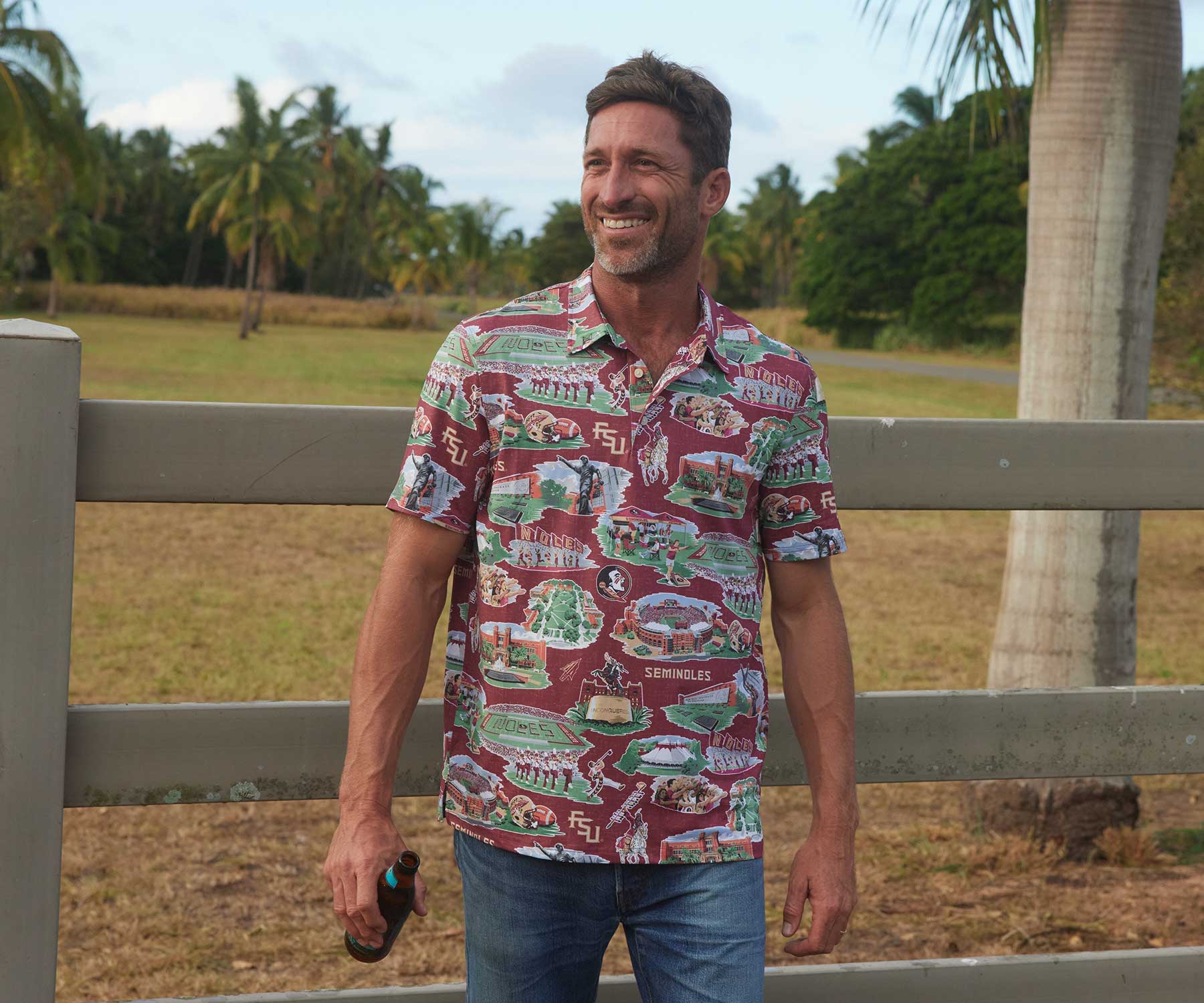 Man in a colorful shirt walking near a fence and palm trees.