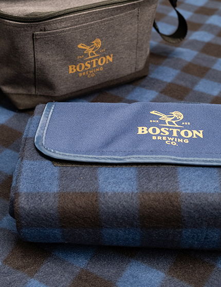 Brown Boston Brewing Co. DTF printed Picnic Rug and Cooler Bag sitting on navy blue plaid rug