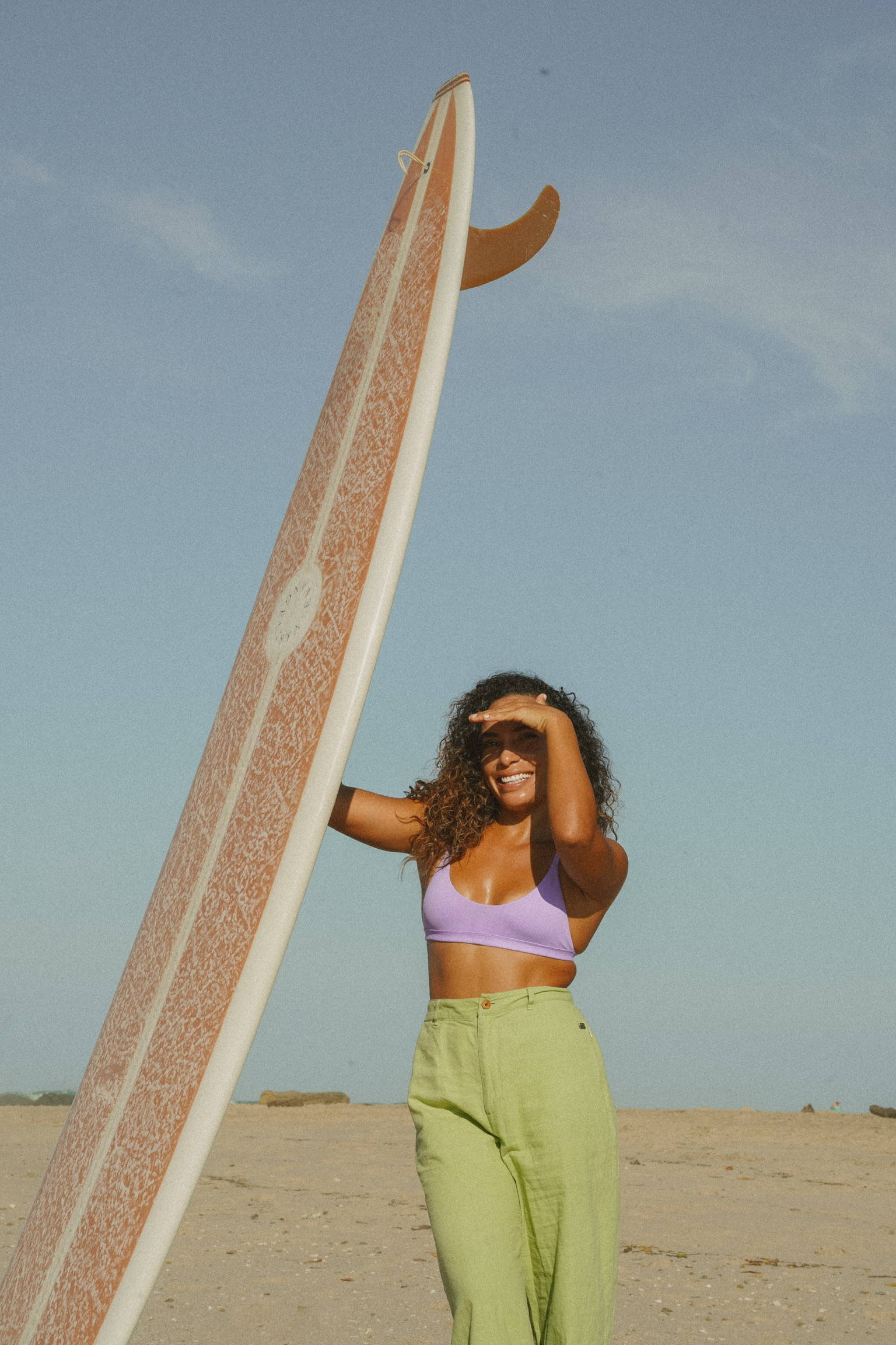 A woman in a lavender bikini top and green pants holding a longboard surfboard on the beach.
