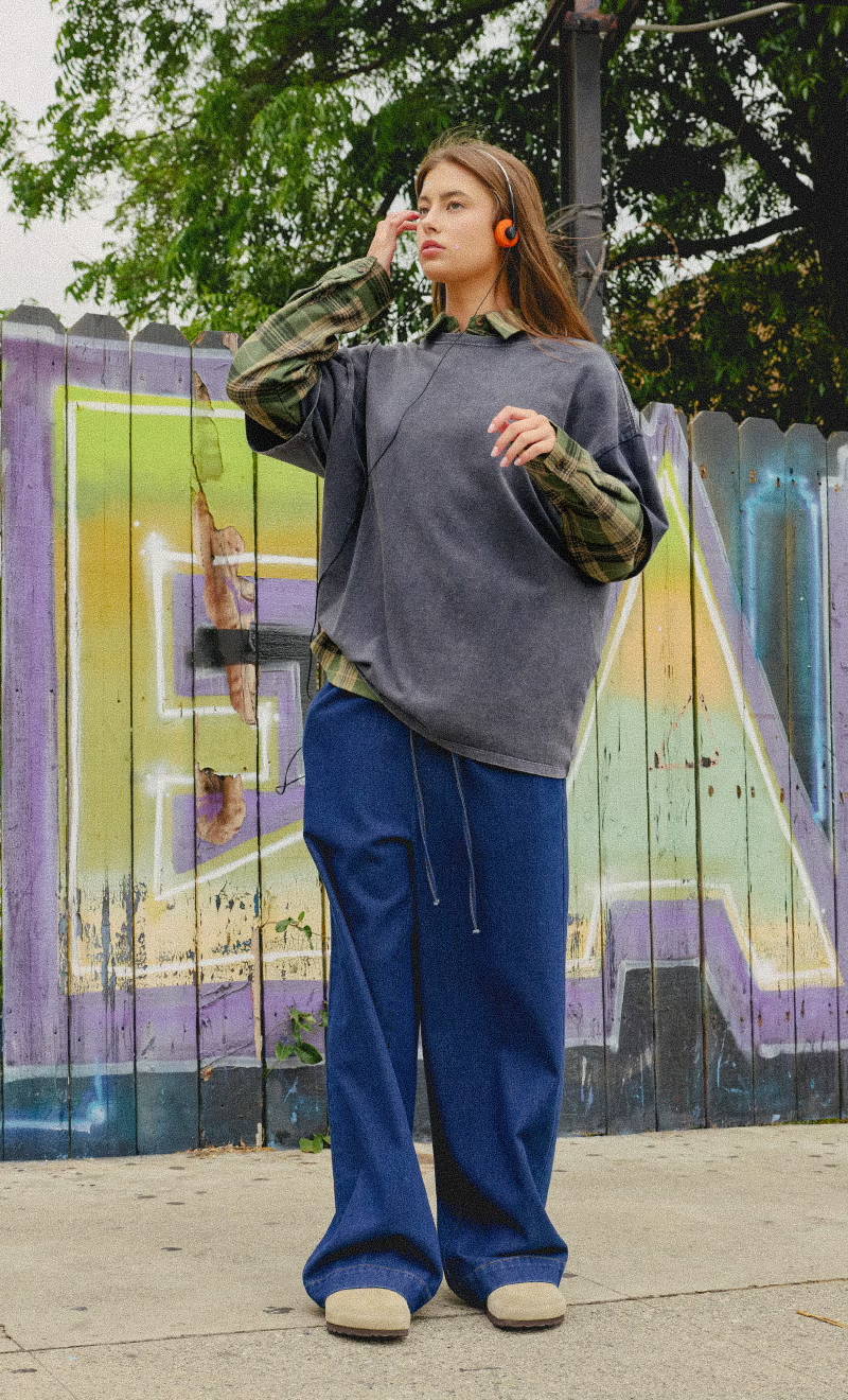 Person wearing a gray oversized shirt and blue pants, standing by a graffiti wall.