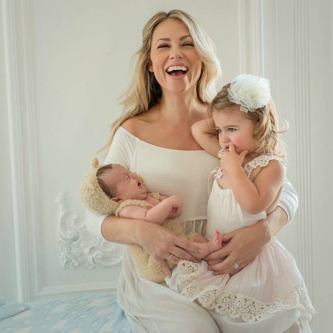 A mom in a white dress seated, holding an infant in a teddy bear onesie and a toddler in a white dress