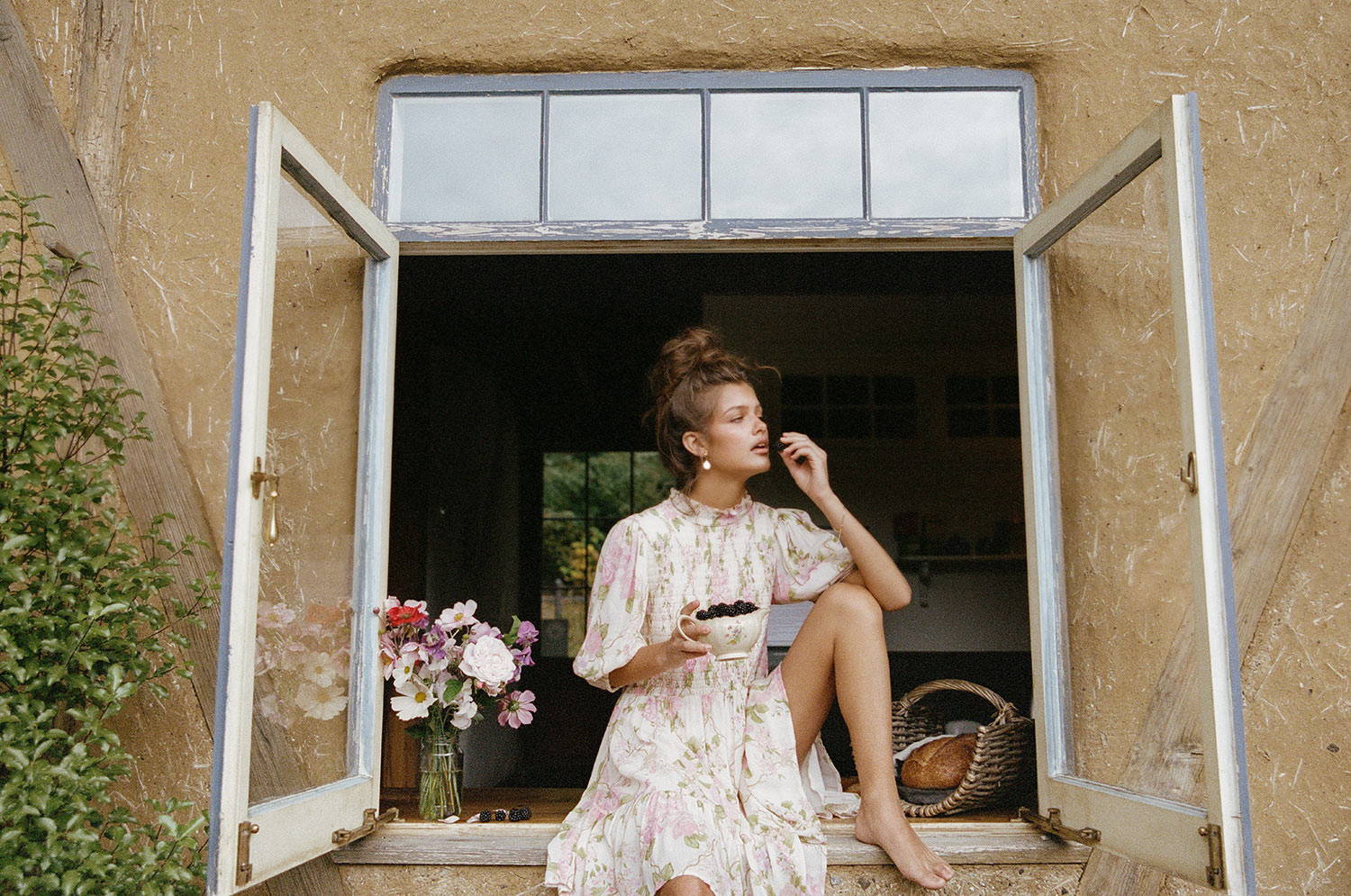 Rose Garden campaign model, Elise Zecevic, sitting on the windowsill of a lake-side cottage while holding a jug filled with mulberries while wearing the rose Rose Garden Mini Dress, featuring high neckline and shirred bodice. 