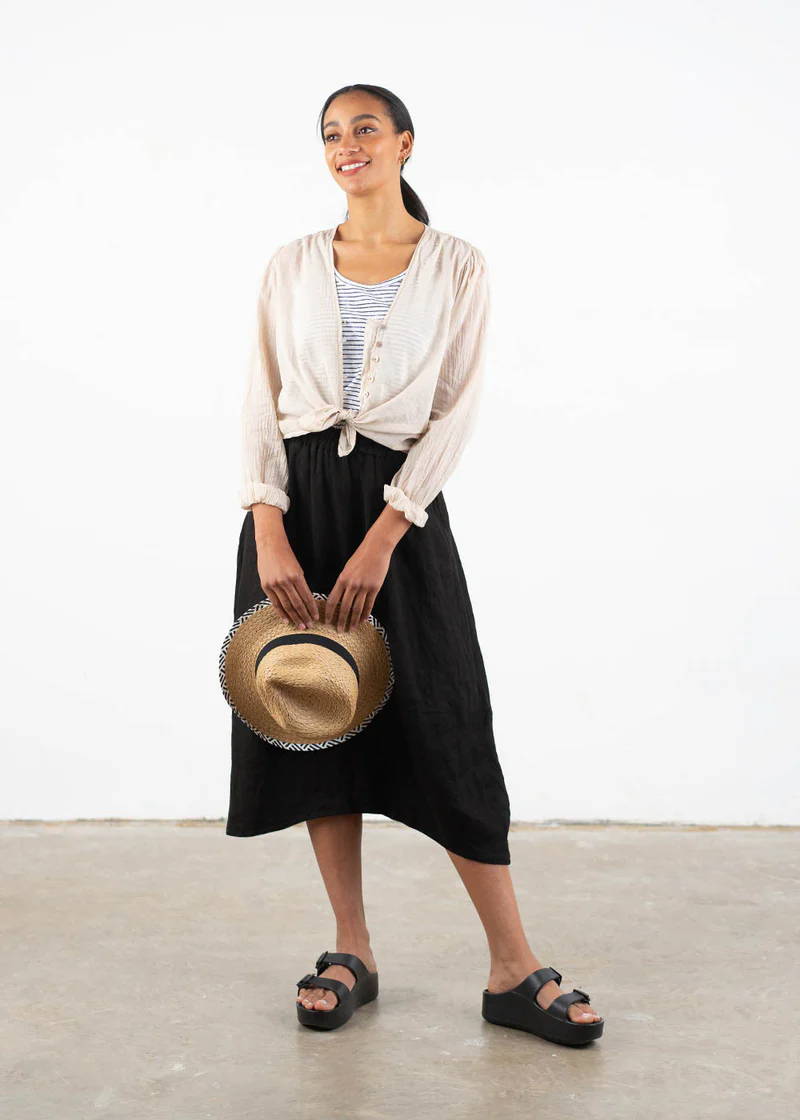 A model wearing a black tulip shaped midi skirt with a oatmeal coloured long sleeved blouse, blue and white striped vest, black chunky platform slides and holding a straw sunhat