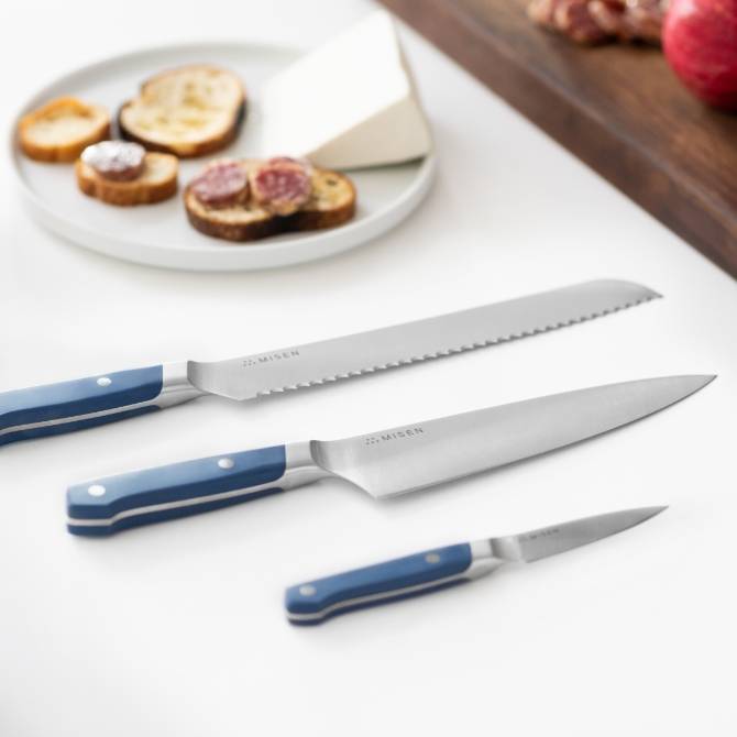 Misen Chef’s, Serrated, and Paring Knives on a white countertop, with a plate of crostini, salami, and cheese in the background.