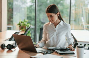 Woman working on a laptop at a desk with plants and stationery around.