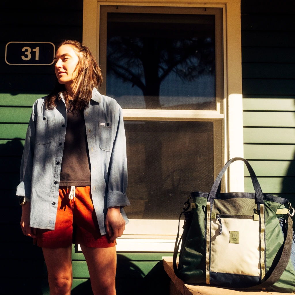 Person standing in front of a house, wearing casual clothing and holding a bag.
