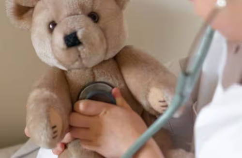 Ein Kind in einem weißen Kittel spielt Arzt. Es hört seinen Teddybären mit einem Stethoskop ab.