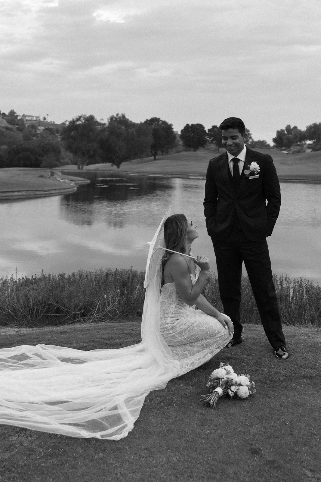 Bride kneeling by groom near a serene lake, dressed in formal wedding attire.