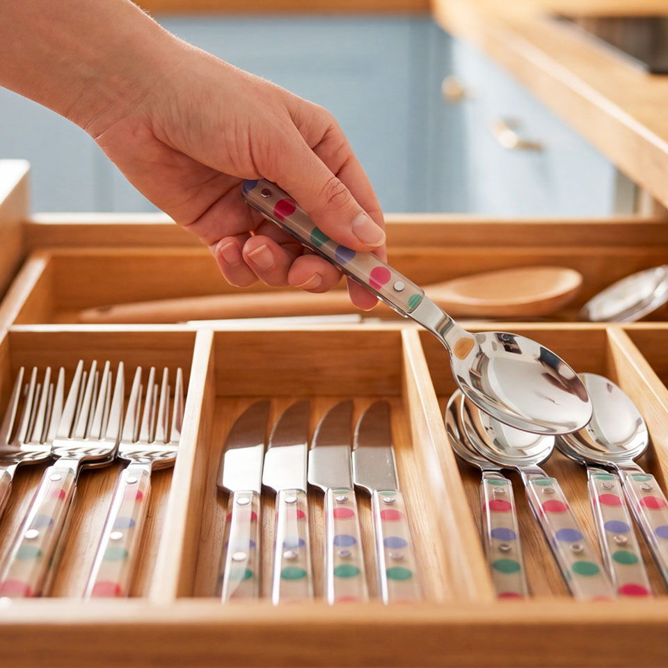 A hand selecting a spoon from a colorful cutlery drawer.