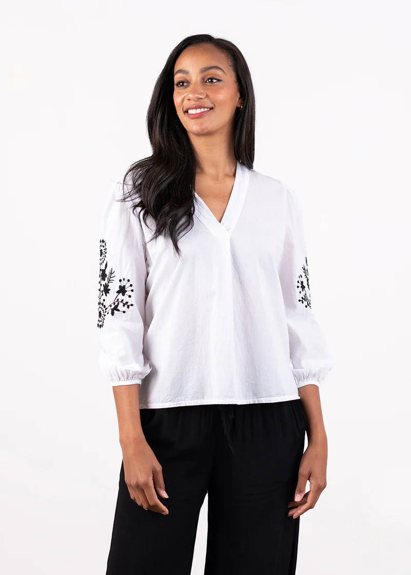 A model wearing a white pull over shirt with black embroidered detailing on the sleeves and elasticated cuffs with black trousers