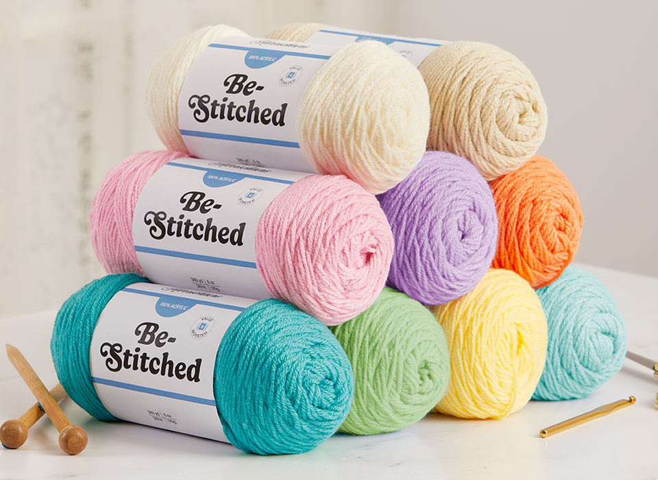 Colorful Herrschners Be-Stitched yarn balls stacked with knitting needles in the background.