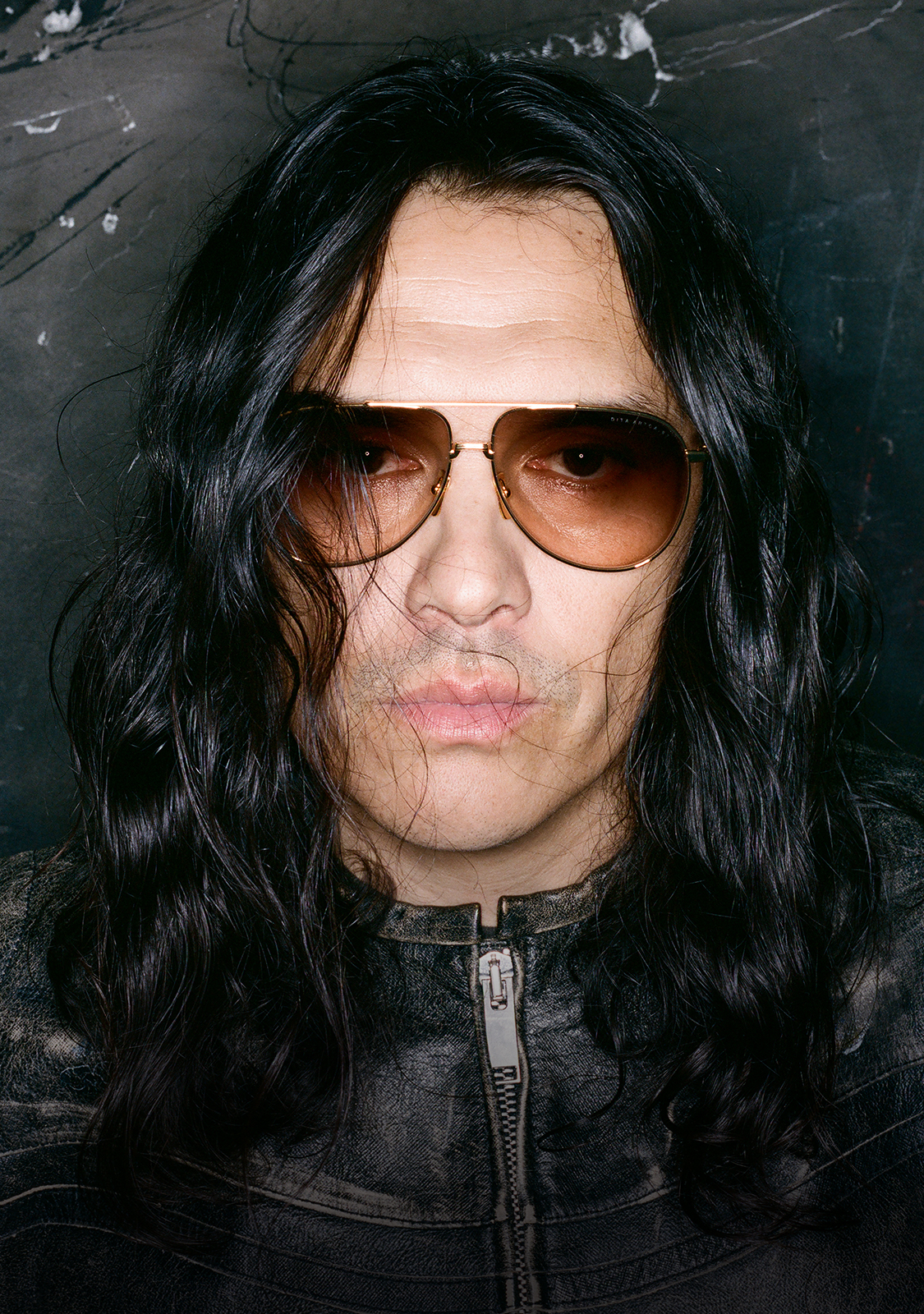 Person with long dark hair and sunglasses, wearing a leather jacket.