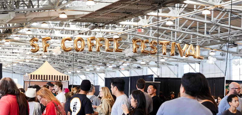 Big Golden Ballon Sing of SF Coffee Festival hanging above the convention center with people milling around all the booths below