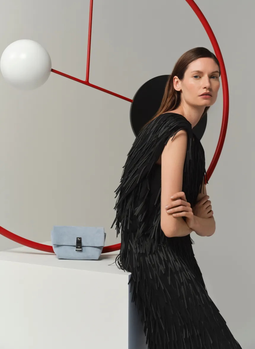 Model in a black fringed dress posing with geometric shapes and a blue handbag.