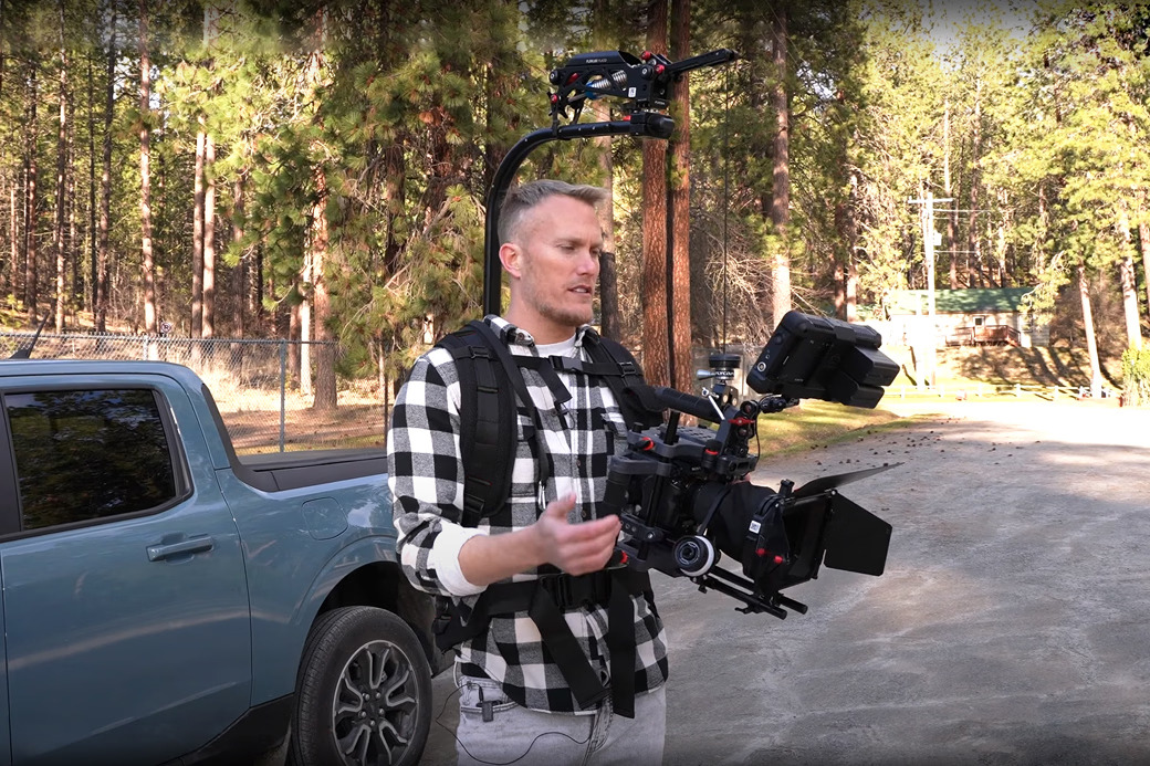 Person holding a camera rig in a forested area near a vehicle.