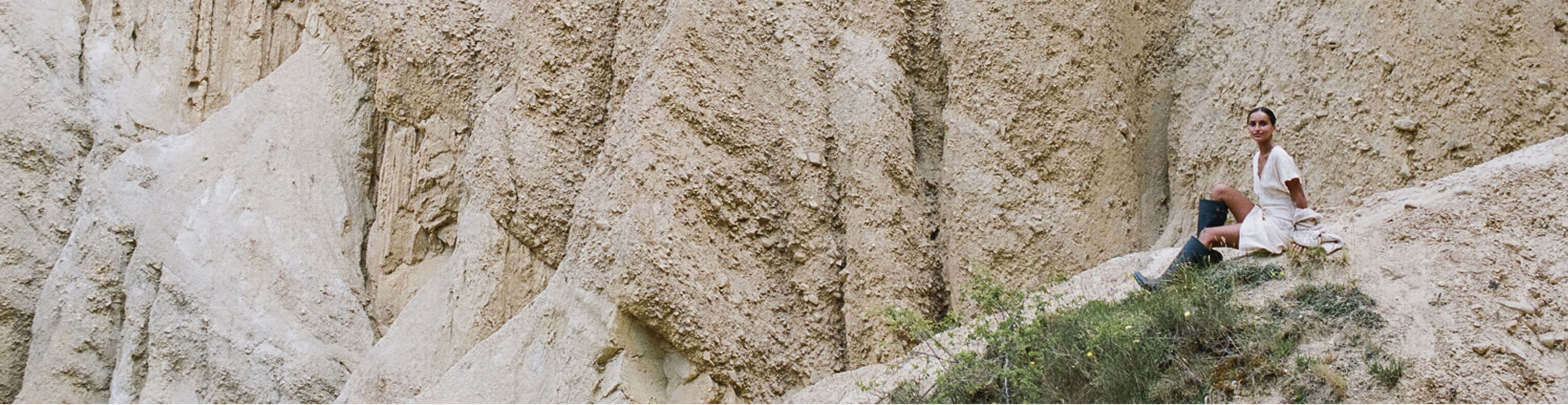 A wide-view image of a sandy-coloured cliff face. Spell Mystic campaign model, Bella Thomas, is posing on a ridge wearing black gum boots and the Petal Jacquard Nightgown. 