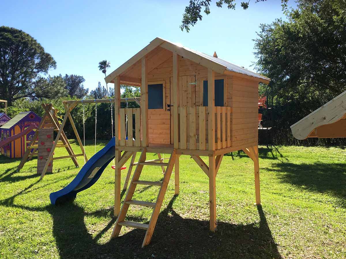Wooden DIY playhouse on stilts with stairs and a blue slide on grass and blue sky in the background by WholeWoodPlayhouses