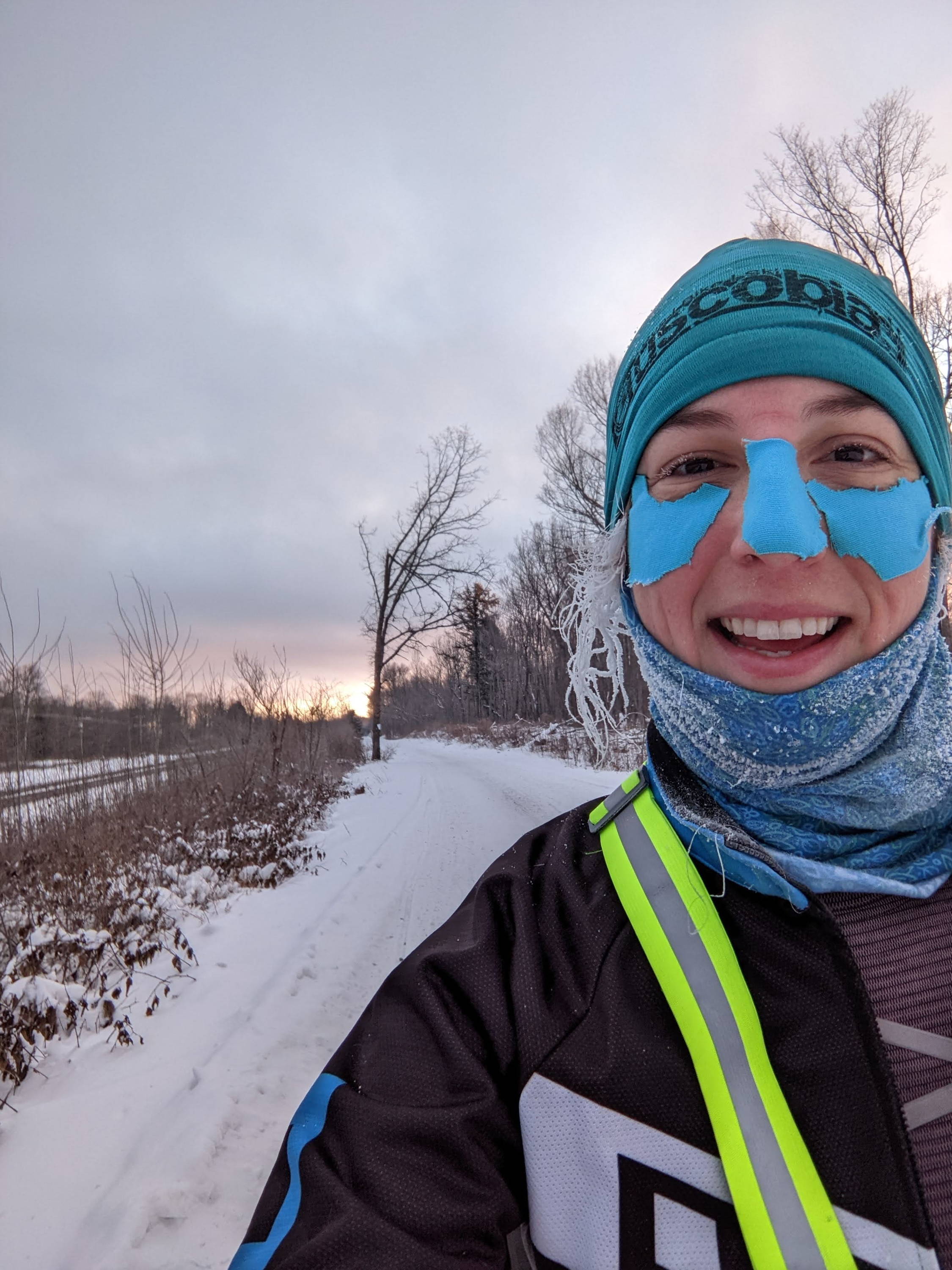 A woman in outdoor gear with frozen hair and face protection smiles in front of a calm sunrise.