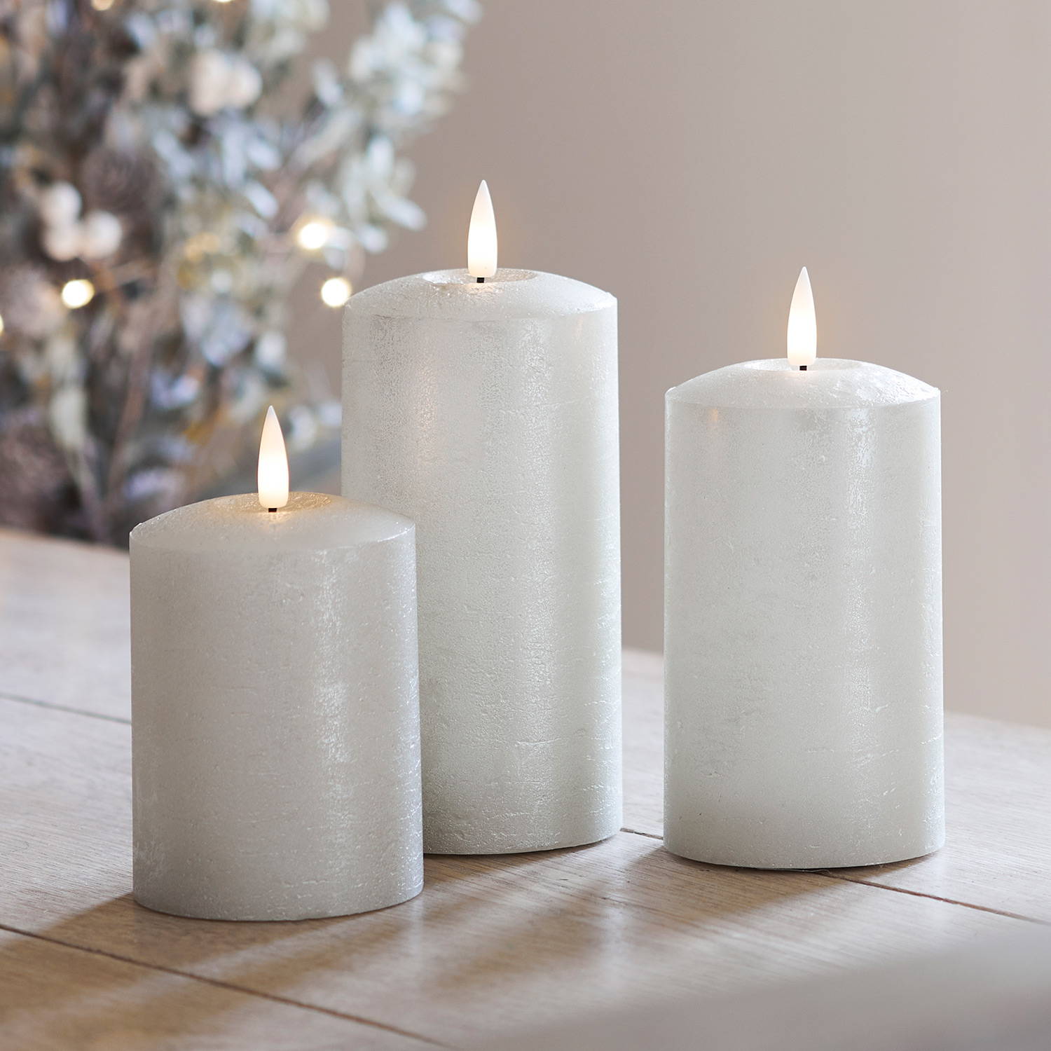 TruGlow® champagne LED pillar candles on a wooden table with a Christmas tree in the background.