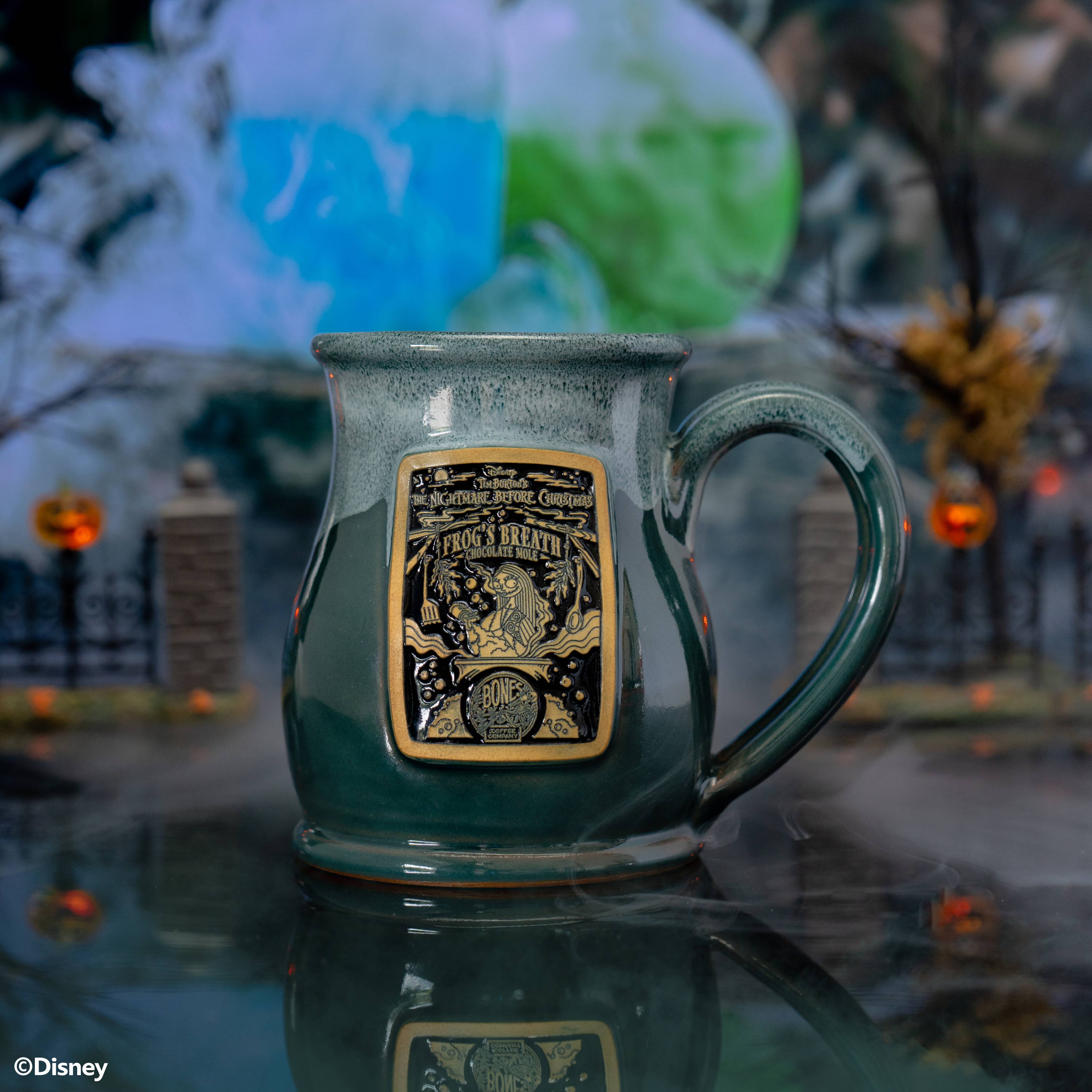 The front of the Bones Coffee Company Frog's Breath hand thrown mug with Sally on the golden medallion. The mug is green with a white glaze on top of it. It is inside a foggy toy graveyard.