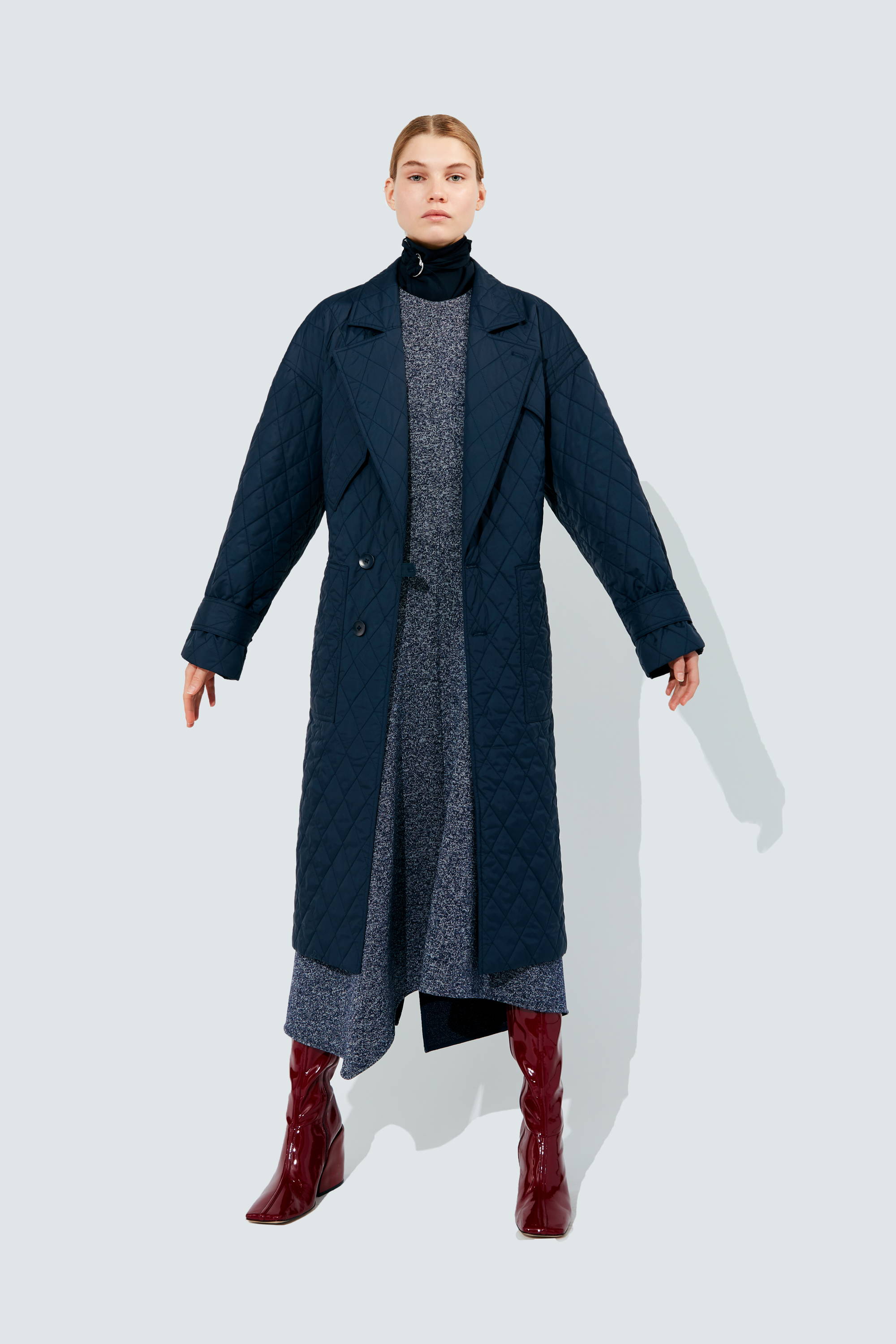 A model wearing a long dark coat and red boots against a light background.