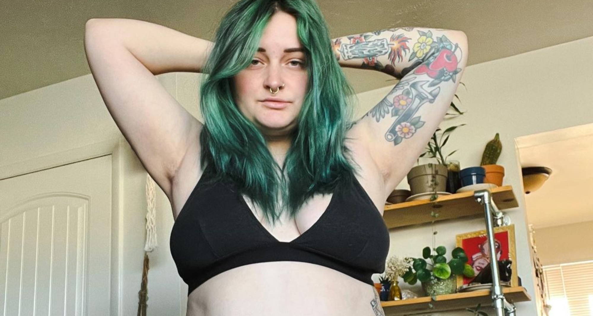 woman with green hair posing in a black triangle bralette with her hands tucked behind her head in front of a shelf of plants