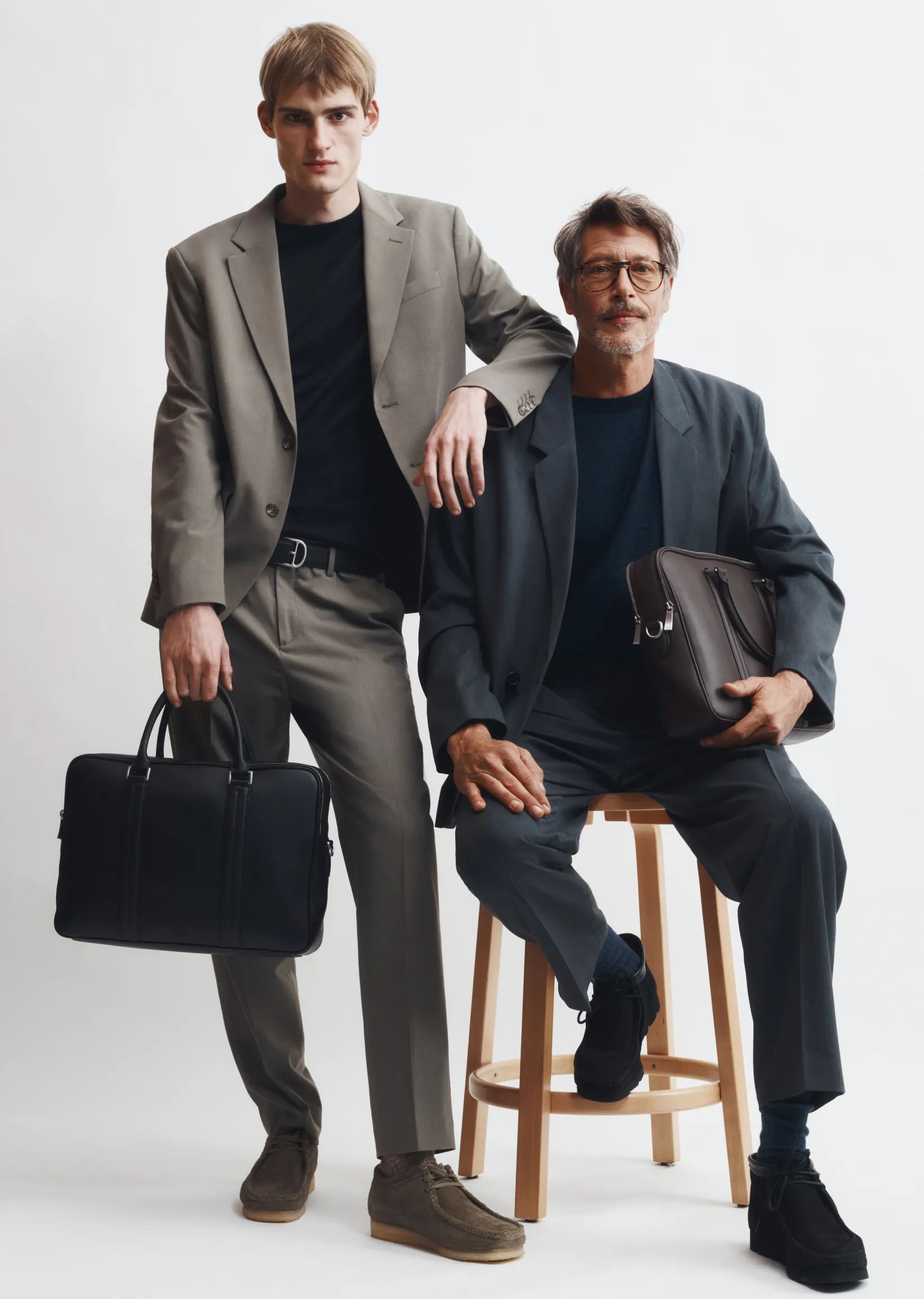 Two men in stylish suits, one sitting on a stool, posing together.