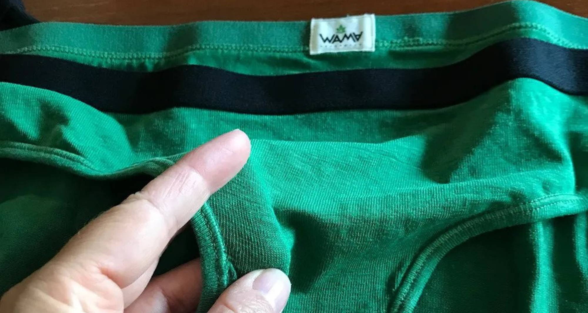 A person holds the gusset of a pair of green hemp underwear by WAMA, laying flat on a brown surface.