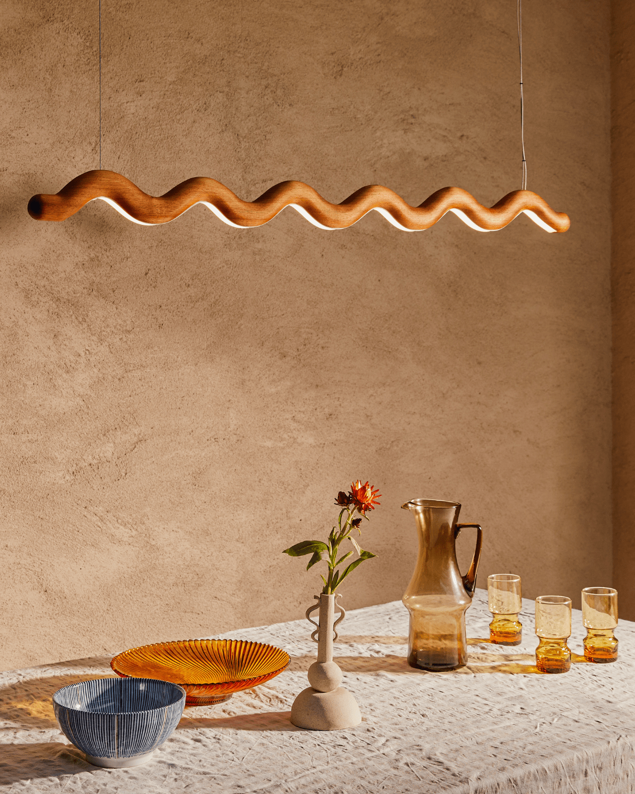 Wavy timber linear pendant light fixture above a dining table with ceramic plates, glasses, and a vase.