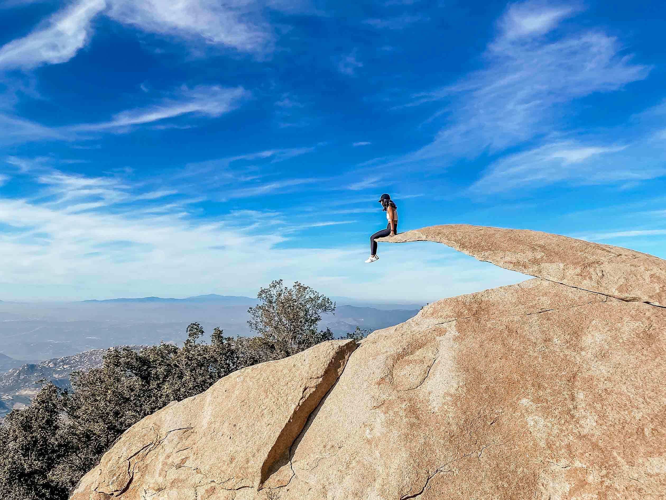 A girl sitting on top of Potato Chip Rock in San Diego, California in a sunny day with some clouds in the distance.