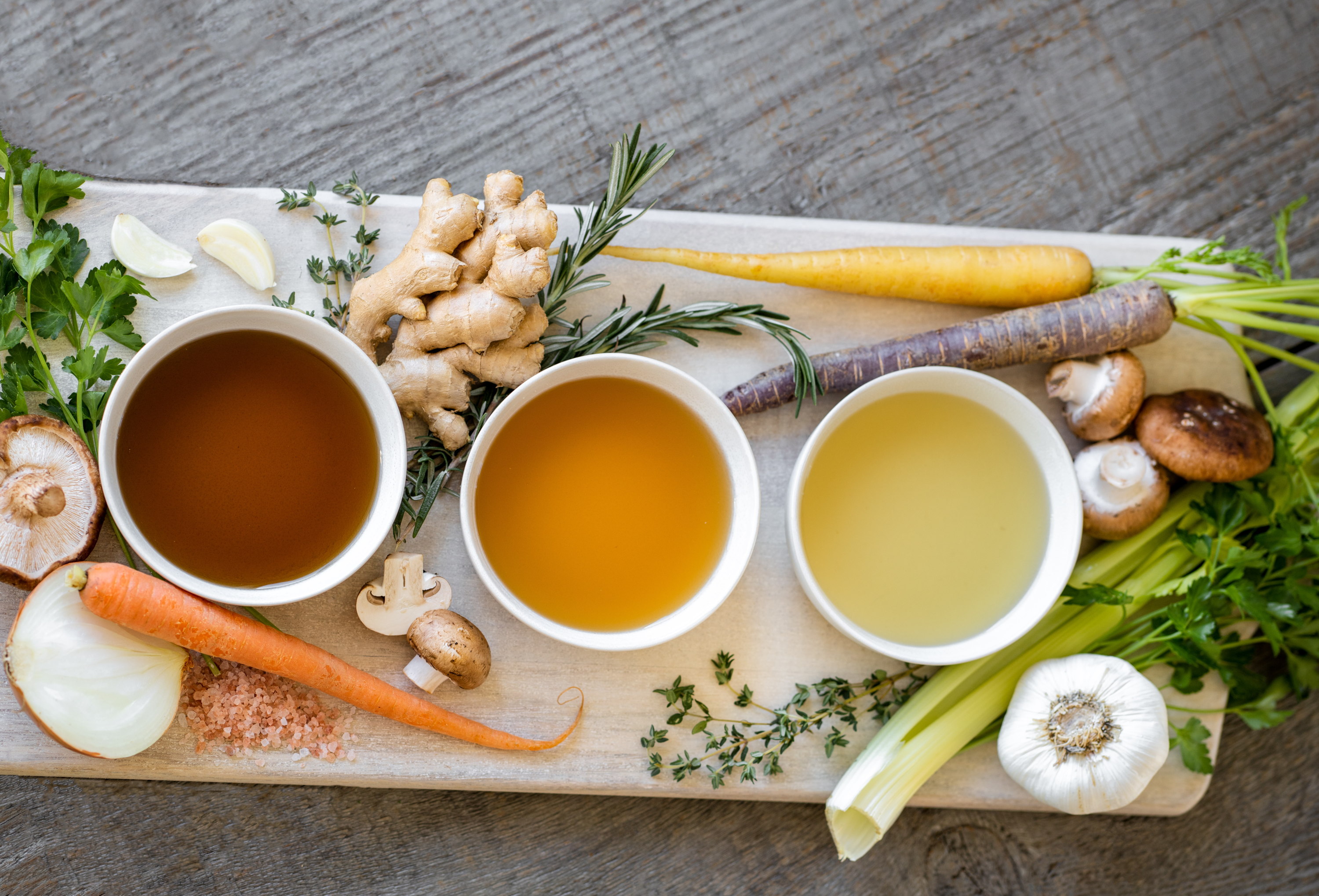 Three different cups of bone broth and a variety of vegetables on a cutting board that aid in immune support.