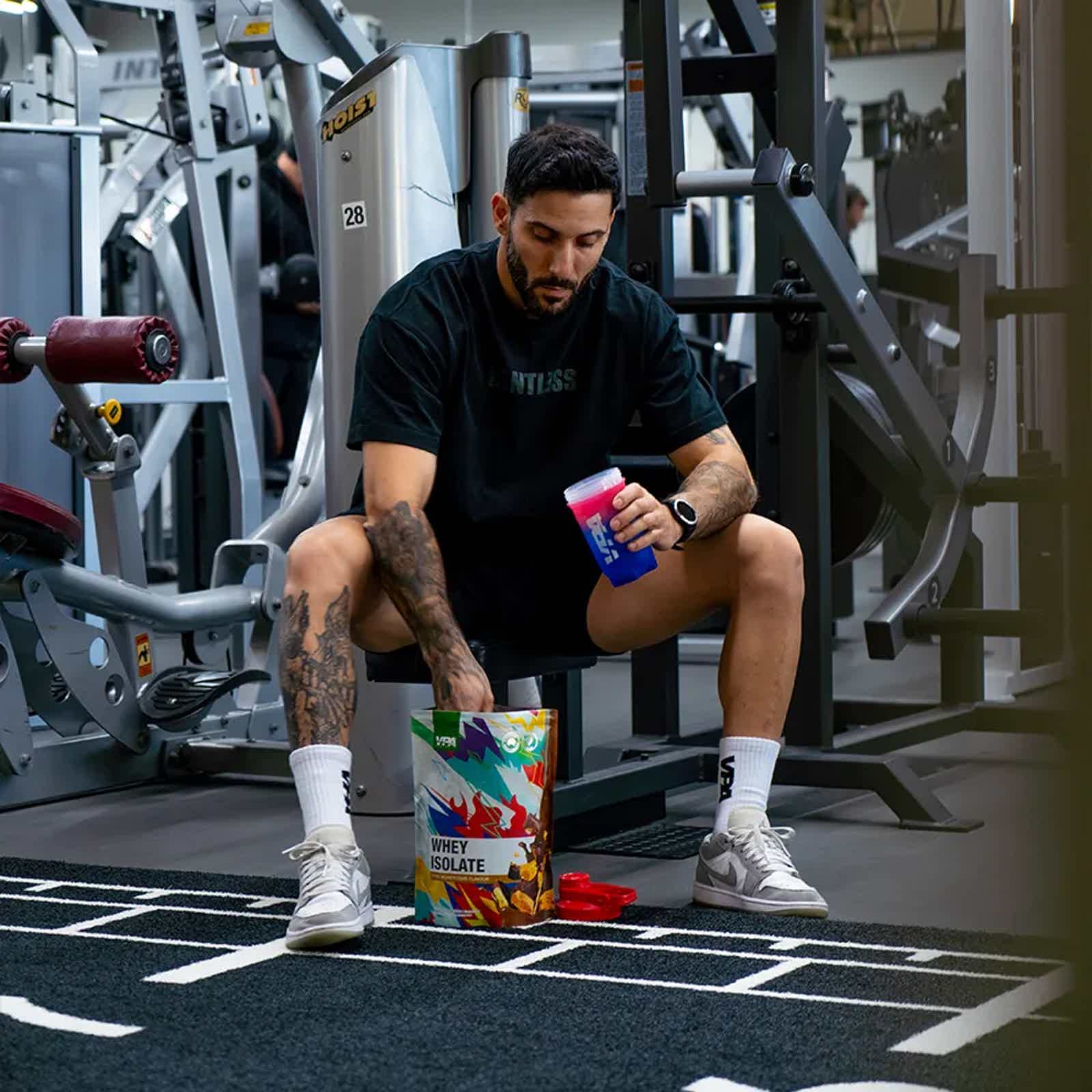 Man sitting in gym, holding shaker and reaching into a colorful supplement container.