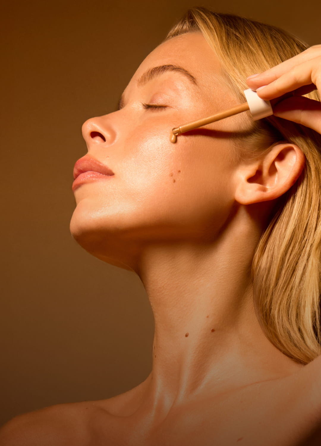 Woman applying facial oil with a dropper, soft lighting and neutral background.