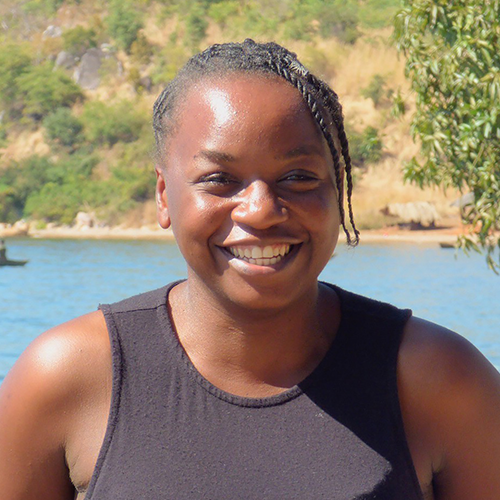 Smiling person with braided hair by a lake in a natural setting.