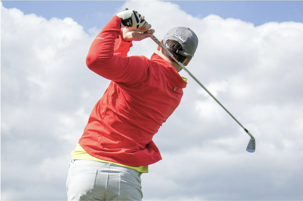 Male golfer viewed from behind mid-swing, wearing a red jacket and grey cap, finishing his shot under a partly cloudy sky.