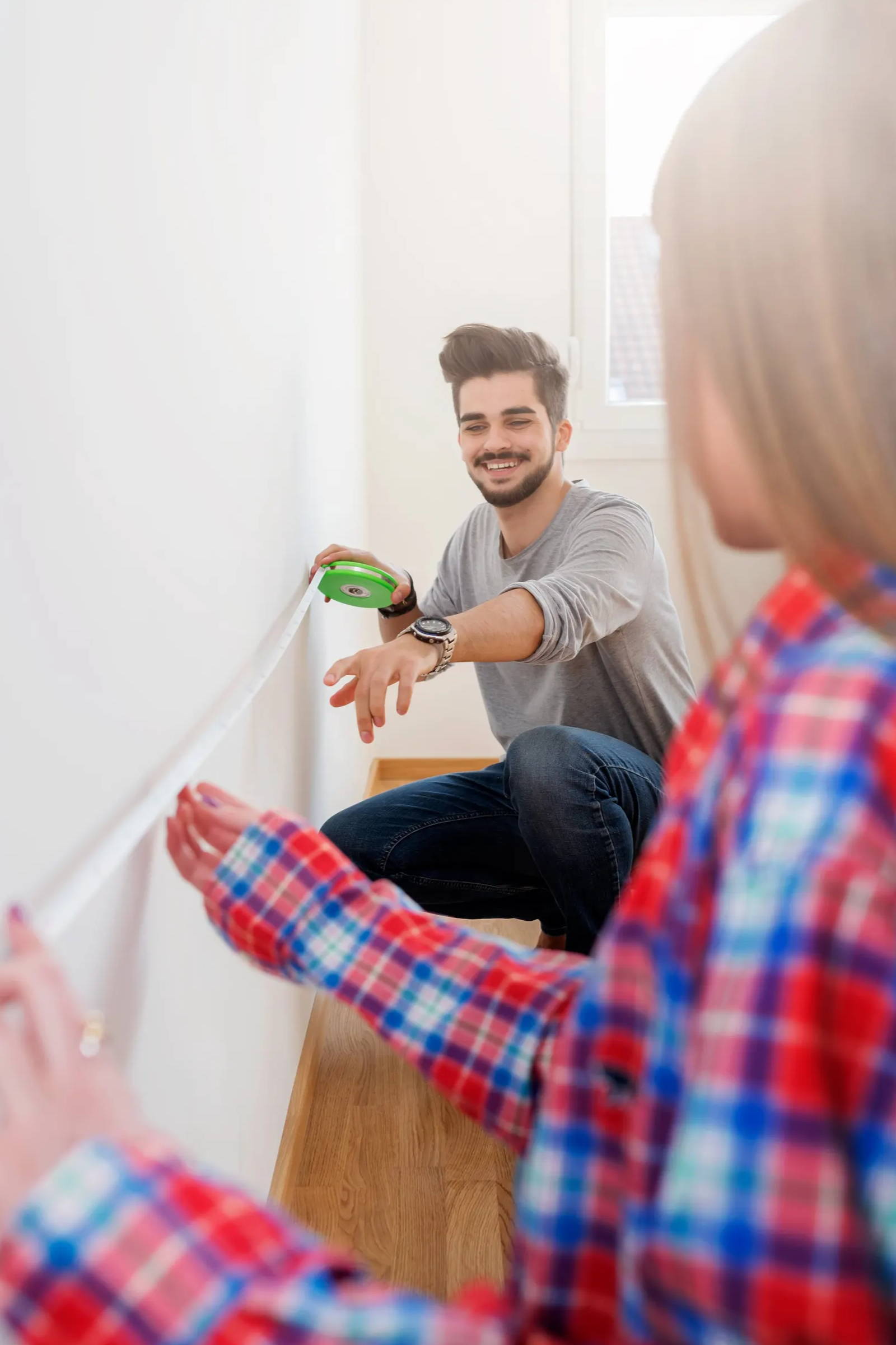 Man smiling measuring wall with extendable measuring tape. Profile of woman's face in foreground.