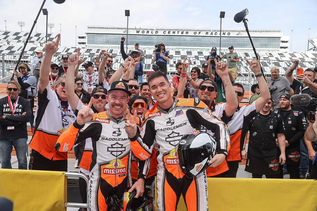 Group celebration photo with racers and fans at Daytona International Speedway, racers pointing upwards in victory.