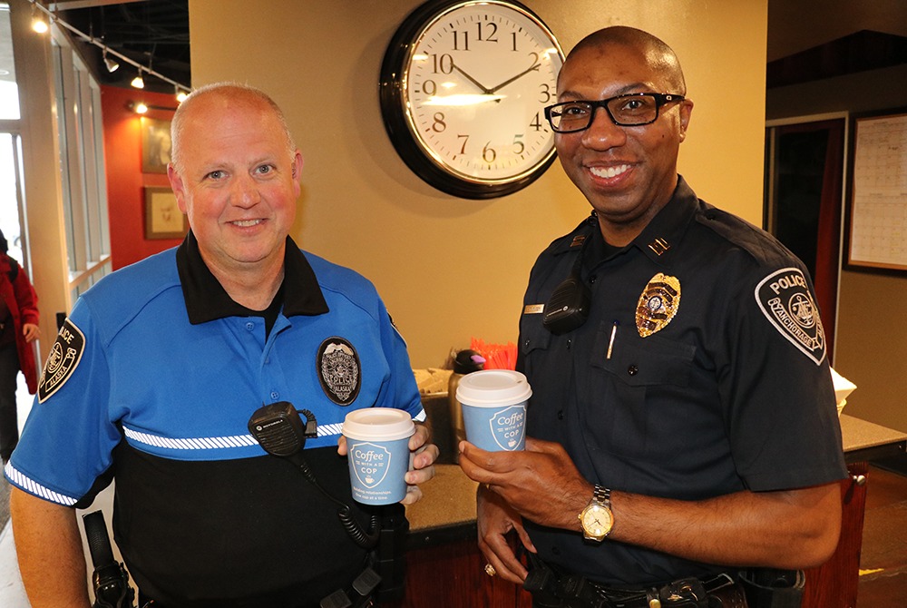 Two police officers holding coffee cups, smiling in a café setting.