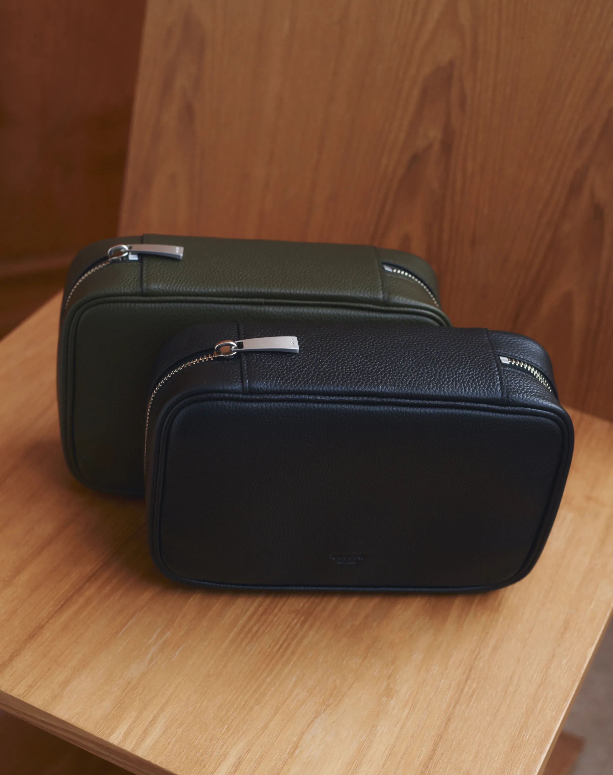 Two leather toiletry bags, one green and one black, on a wooden surface.