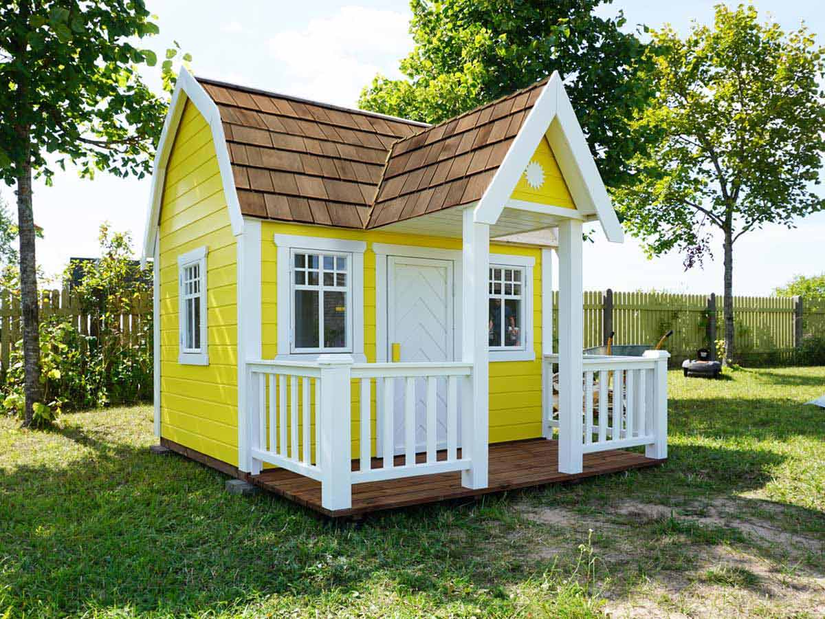 Custom playhouse in yellow color with shingles roof in a green garden by WholeWoodPlayhouses