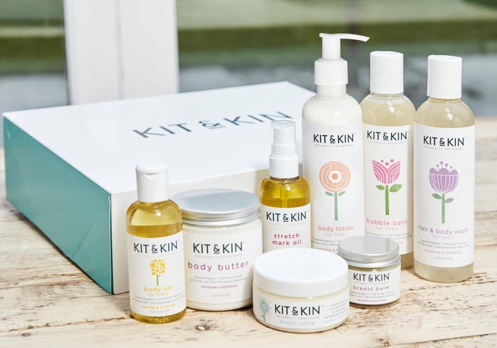 A collection of Kit and Kin skincare products in plastic containers are grouped in front of a gift box.