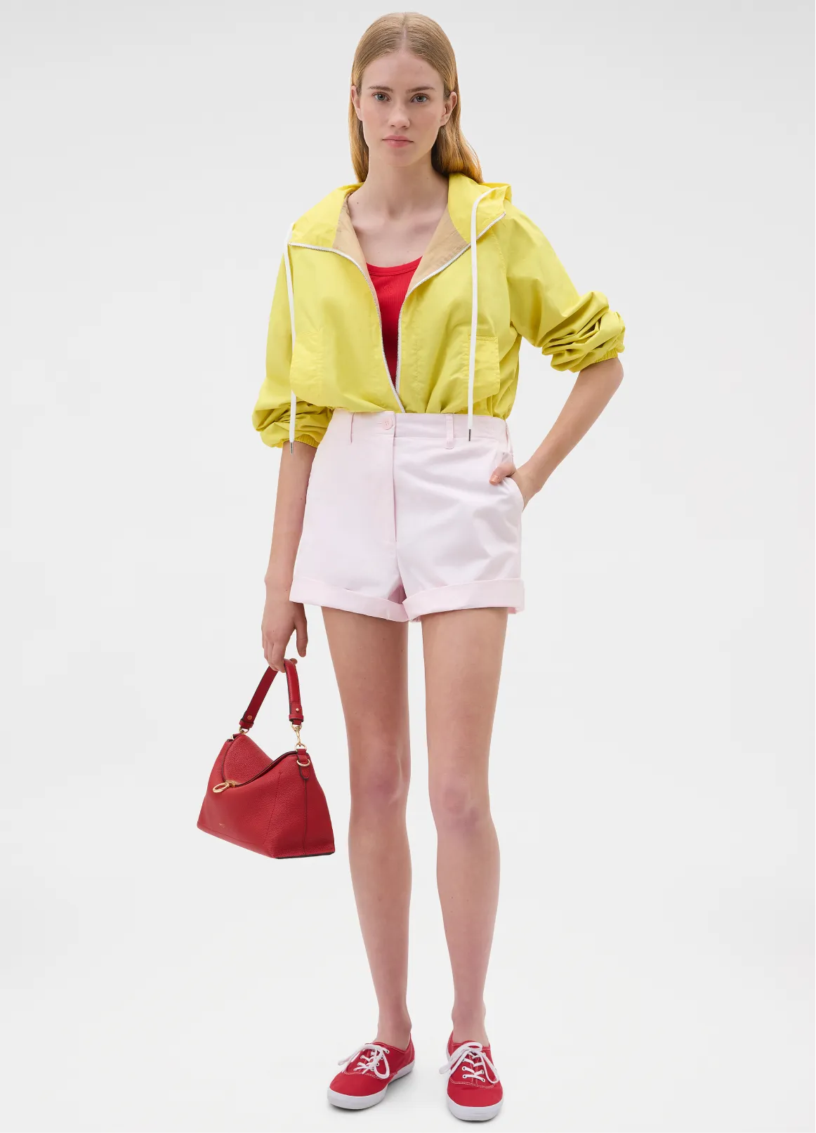 Model wearing yellow jacket, red top, pink shorts, holding red bag.