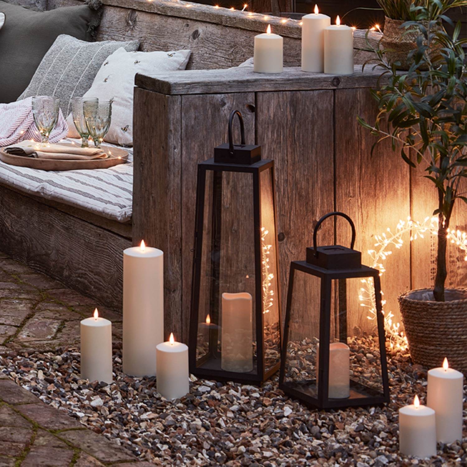 2 Black lanterns on the floor of an outdoor seating area with LED candles and a  starburt light in the background.