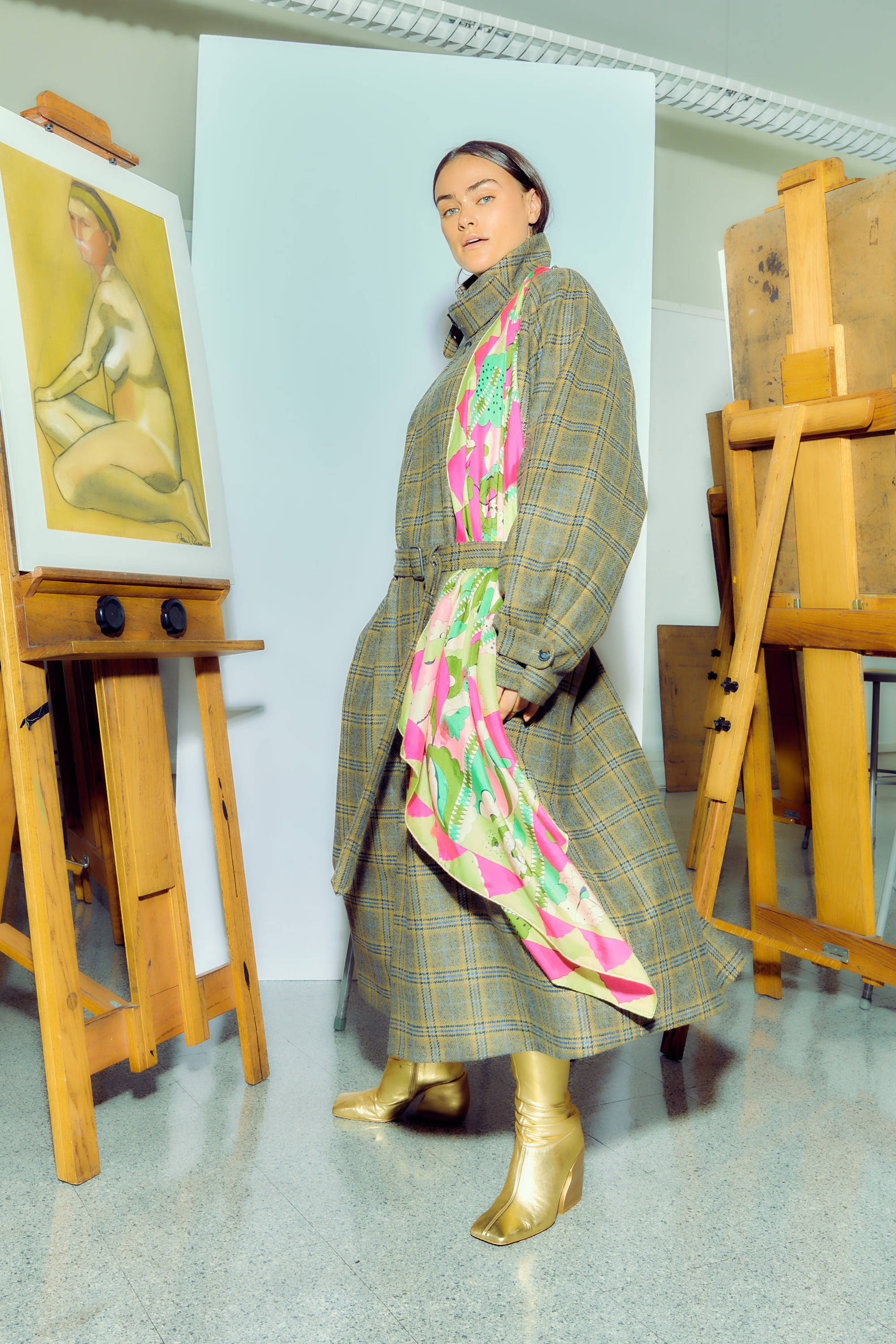 Model in stylish outfit poses beside art easels and a painting.