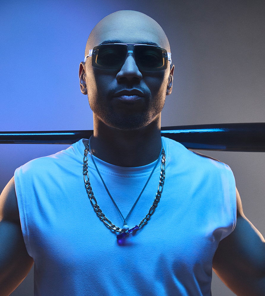 A man in sunglasses with a bald head and a chain, set against a blue background.