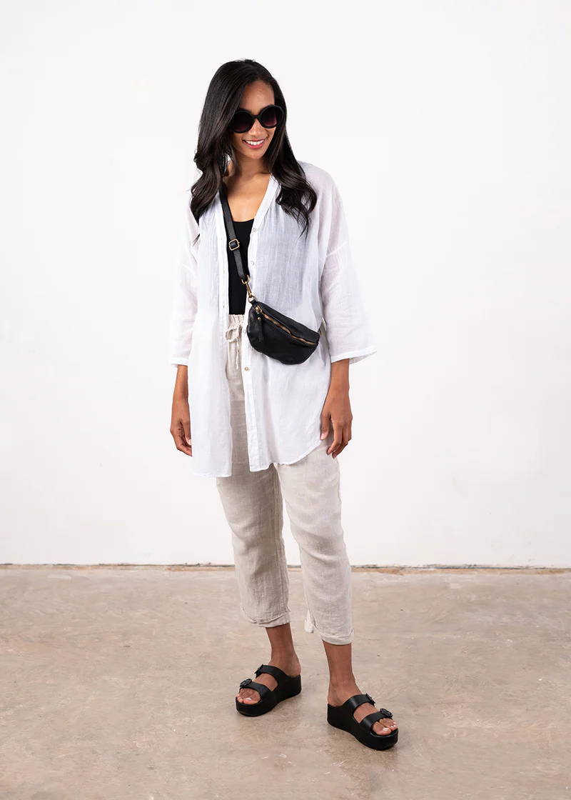 A model wearing a white semi sheer shirt over a black vest, off white trousers, black chunky slides with a black cross body bum bug and black sunglasses